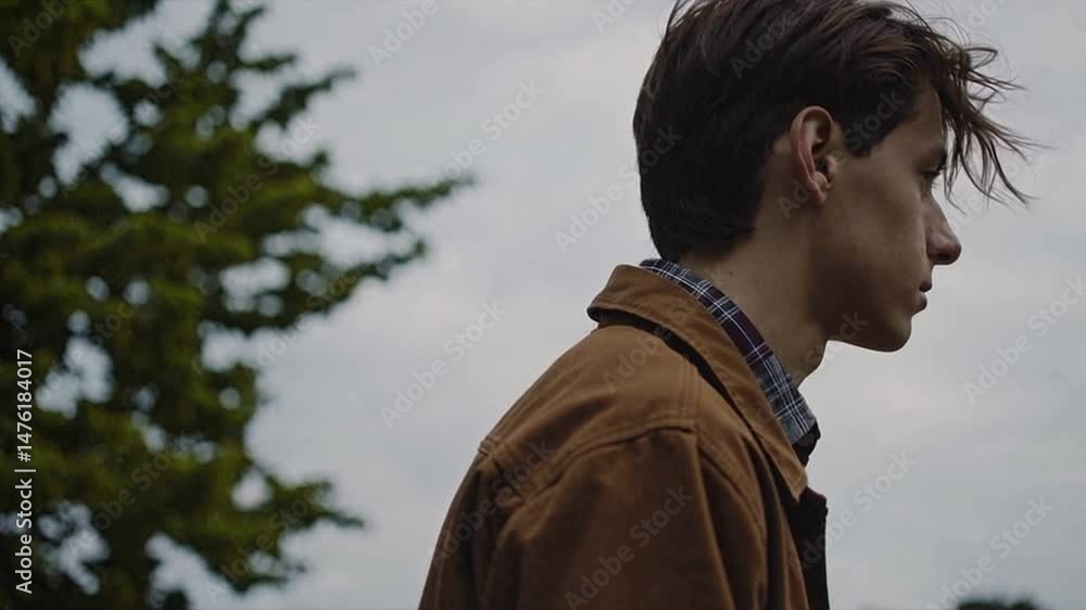 Profile of a young Caucasian male with brown hair, wearing a brown jacket and plaid shirt. He stands outdoors against a cloudy sky, surrounded by greenery. Profile of a young Caucasian male with brown hair, wearing a brown jacket and plaid shirt. He stands outdoors against a cloudy sky, surrounded by greenery.