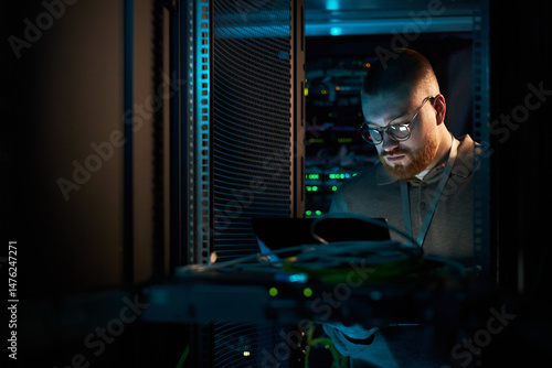 Technician in server room inspecting hardware components of data center equipment, surrounded by servers and cables, engrossed in work while holding tablet for diagnostics