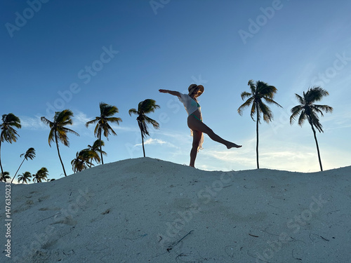 Woman walking through the sand dunes at sunset with palm trees in the background. A tranquil and contemplative moment as she strolls along the golden sands, bathed in the warm hues of the setting sun.