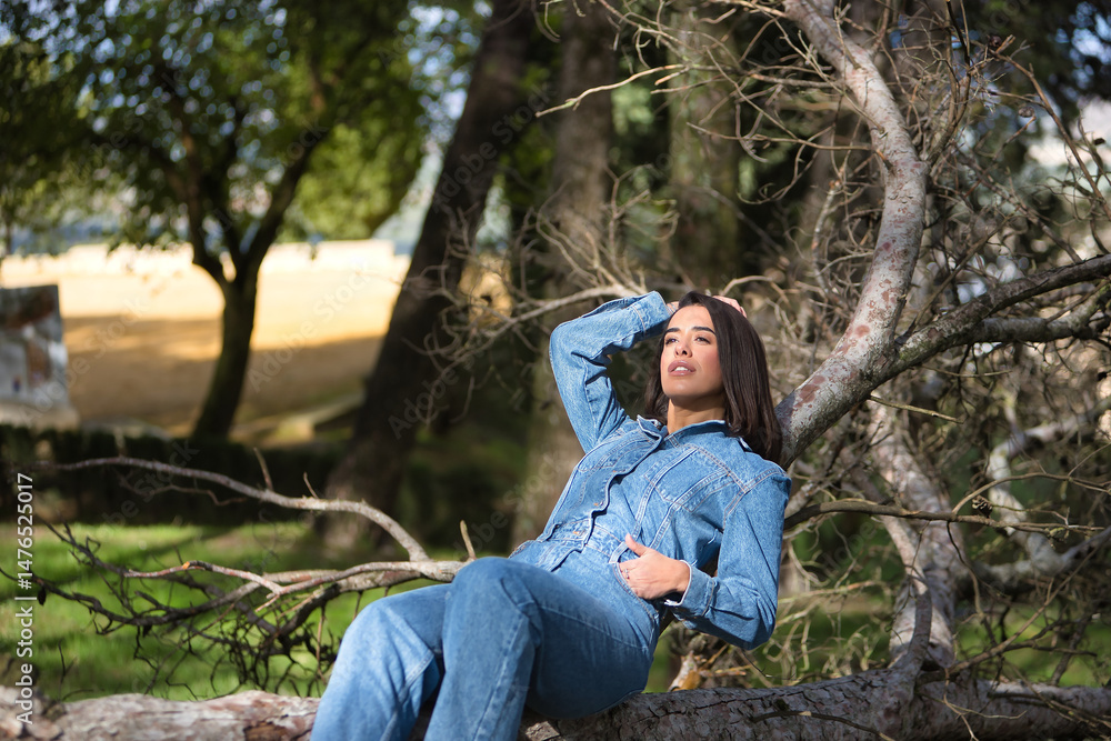 Young, beautiful, brunette, Hispanic woman, wearing a denim suit, pensive and distracted, lying on the fallen trunk of a tree, in nature, receiving the sun's rays. Concept fashion, beauty, femininity. Young, beautiful, brunette, Hispanic woman, wearing a denim suit, pensive and distracted, lying on the fallen trunk of a tree, in nature, receiving the sun's rays. Concept fashion, beauty, femininity.