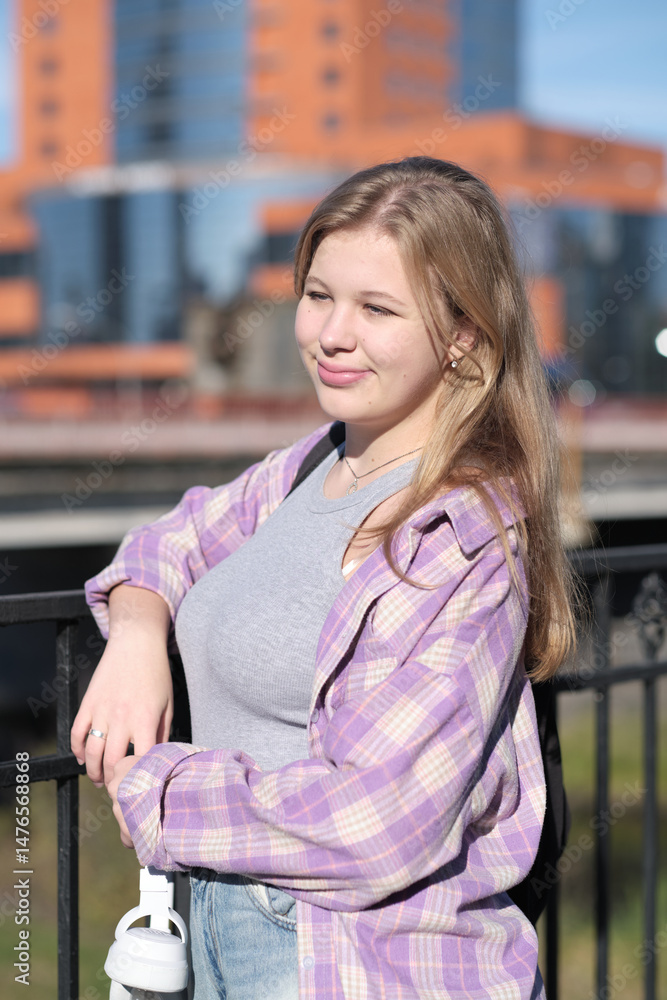Fototapeta premium A teenage girl enjoys the sunlight as she poses by a railing, holding headphones in her hand. The photo captures the vibrancy and freedom of youth in an urban setting.