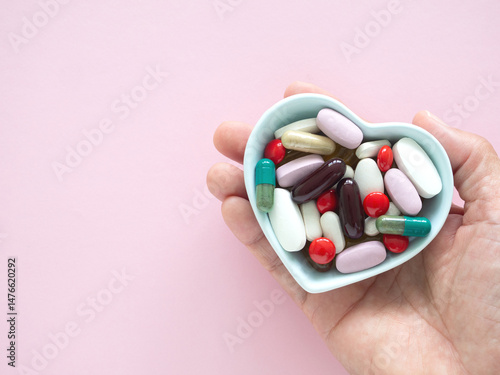 Hand holding a heart-shaped blue bowl filled with various colorful pills and capsules over pink background. Concept daily routines and medication management supplements..