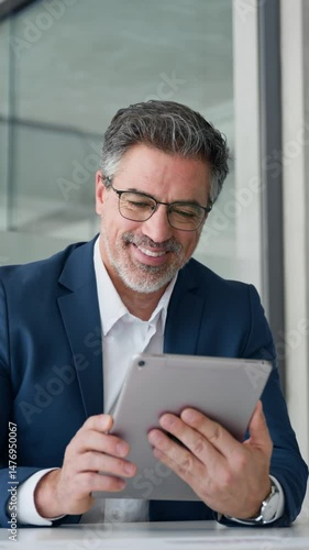 Senior businessman entrepreneur sitting at desk working on financial sales project, looking smiling at screen. Mature 50s Latin business man ceo manager using tablet pad computer in office. Vertical