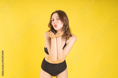 Wallpaper Mural Confident young brunette woman in black bikini blowing a kiss while smiling in studio with yellow background, expressing fun playful energy for beauty, fashion, or lifestyle concepts Torontodigital.ca