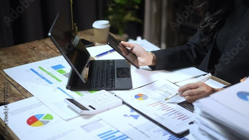 A woman is sitting at a desk with a laptop and a tablet. She is using the tablet to look at a document on the laptop