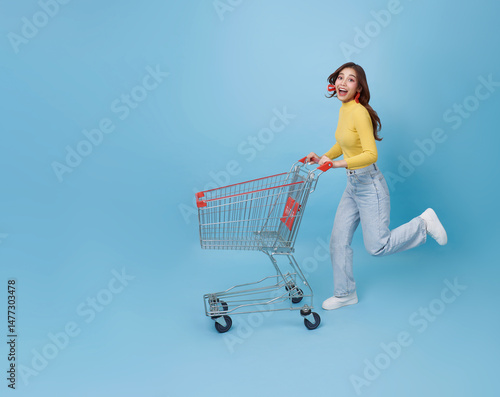 Happy excited Asian teen woman pushing empty shopping cart or shopping trolley running promotion supermarket isolated on blue background.