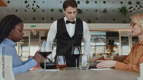 Side view shot of young Caucasian waiter in classic outfit listening respectfully to middle-aged female guest complaining about ordered dish while having lunch with her Black girlfriend in restaurant