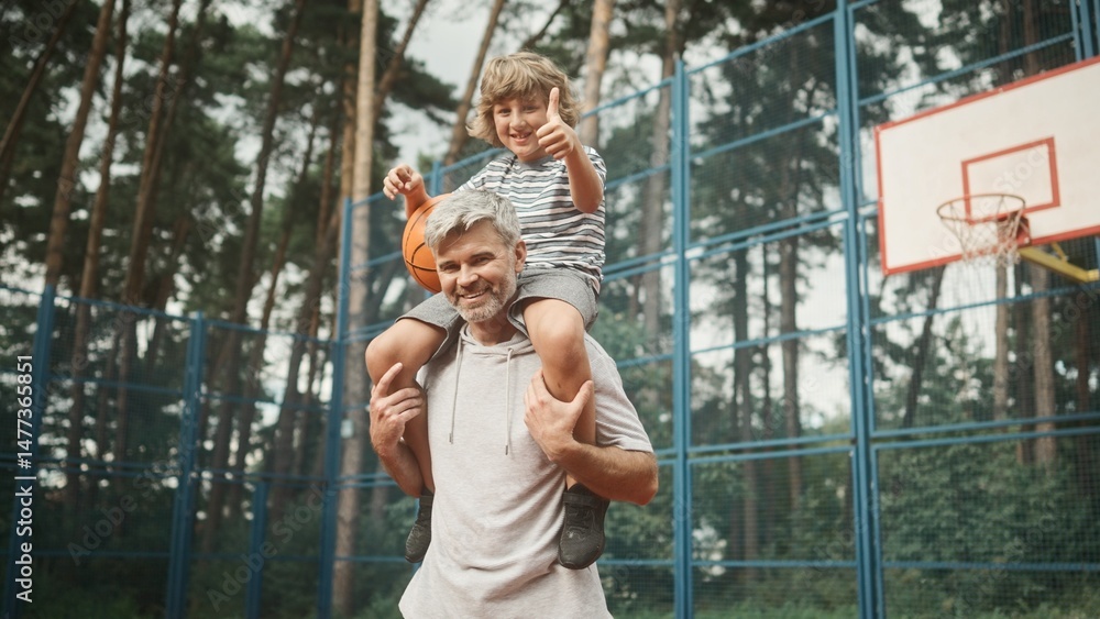 Obraz premium ?ute little boy is sitting on dad's shoulders and holding basketball. Loving father and son smiling, posing and looking at camera on background of basketball court. Family spending time together.