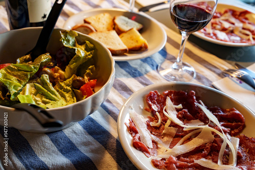 Carne salada (EN: Salted meat) a traditional meat dish from Trentino. Beef served with olive oil, and Parmesan shavings. On a set table with mixed salad, toasted bread, and a glass of red wine