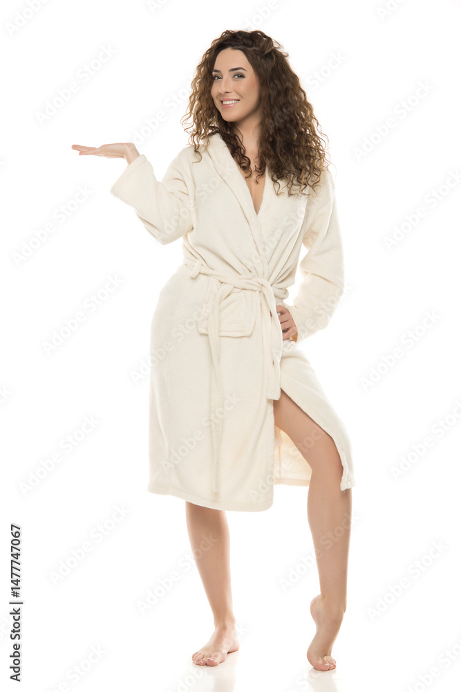 Smiling woman with curly brown hair in white bathrobe presenting invisible product against white background, advertisement pose, marketing concept Smiling woman with curly brown hair in white bathrobe presenting invisible product against white background, advertisement pose, marketing concept