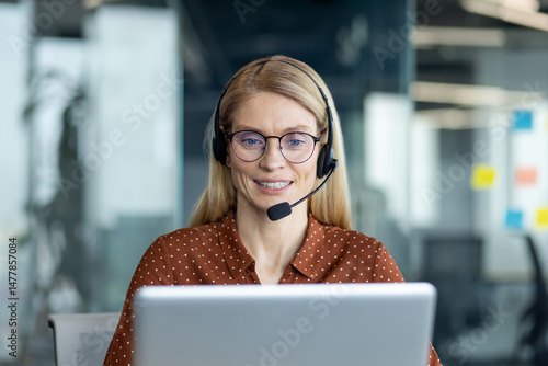 A friendly customer service representative smiles while wearing a headset and working on a laptop at a modern office.