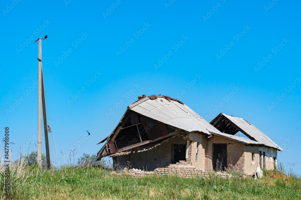 Obraz premium A close-up of two dilapidated one-story brick houses and a pole without wires, standing in the middle of a meadow against a blue sky. A common Roller or raksha (Coracias garrulus) bird hunts nearby.