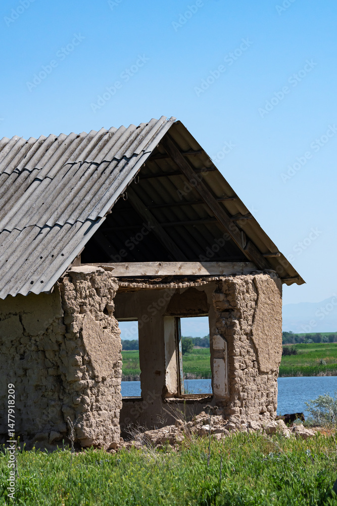 A vertical close-up photo of a destroyed one-story brick house with no windows or doors, with empty openings, against a blue sky; a river is visible through the windows. A vertical close-up photo of a destroyed one-story brick house with no windows or doors, with empty openings, against a blue sky; a river is visible through the windows.