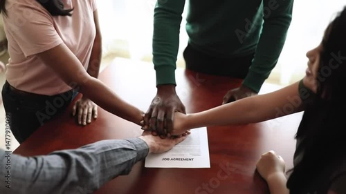 Diverse group placing hands above job agreement on wooden table in office space