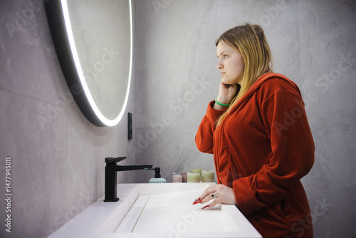 Εκτύπωση καμβά Young woman in a red bathrobe leans against the sink, resting her face on her hand with a weary expression while staring into a glowing round mirror