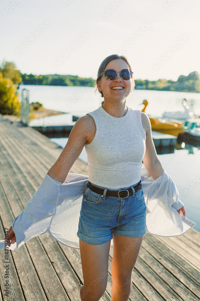 Cheerful woman in shorts and sunglasses walking along the pier near the lake. Woman having fun and running under the scorching sun. Young woman on a walk near the lake.