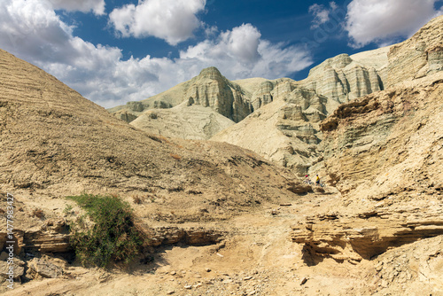 Breathtaking arid landscape featuring rugged beige rock formations and eroded cliffs under a vivid blue sky with scattered clouds. The scene includes two cyclists navigating through the canyon, adding