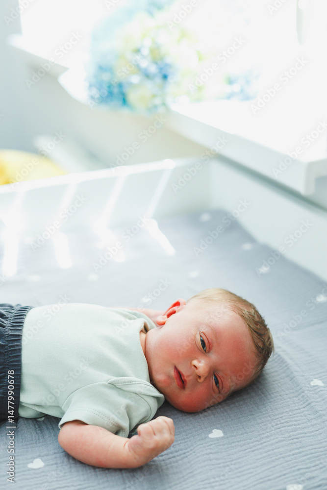 An adorable little baby is seen relaxing peacefully on a soft gray mattress positioned near a window, with beautiful natural light gently streaming in, creating a serene and warm atmosphere