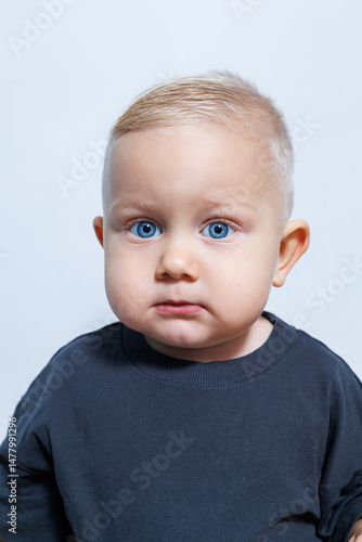 Obraz na płótnie This charming little baby, sporting captivating blue eyes and light blonde hair, is dressed in a casual outfit, positioned against a calm and neutral background that enhances their cuteness w salonie