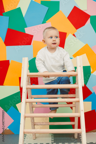 Obraz na płótnie A toddler enthusiastically climbs a wooden ladder set against a vibrant backdrop filled with colorful geometric shapes, showcasing the joy of childhood exploration and adventurous play w salonie