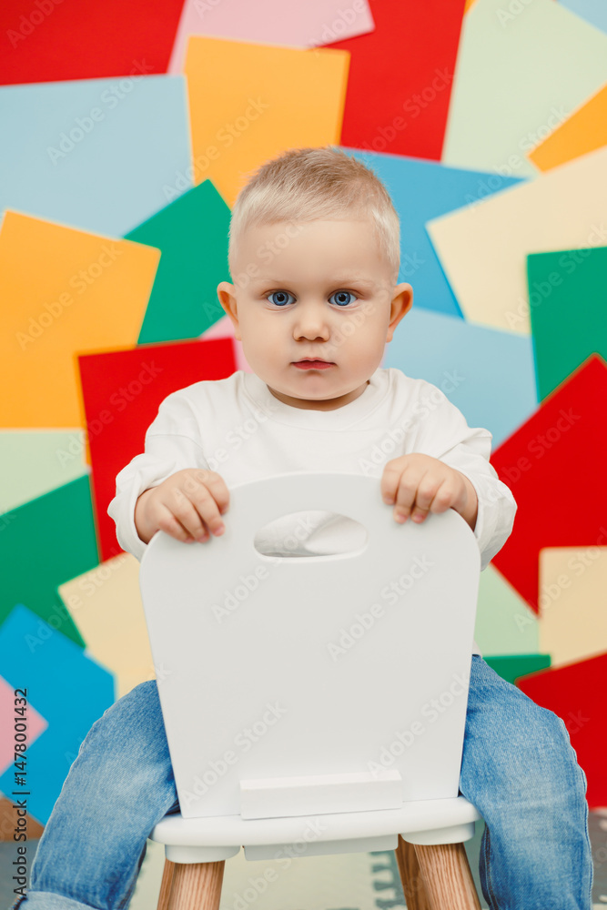 An adorable and cheerful toddler is sitting contently on a chair, against a vibrant and colorful background that enhances the artistic atmosphere of this candid photography moment