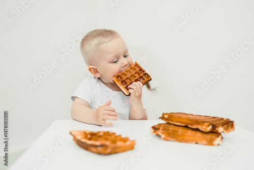 Obraz na płótnie A delightful toddler is seen happily enjoying a delicious waffle treat while sitting at a minimalist white table, showcasing a joyful moment filled with sweetness during breakfast time w salonie