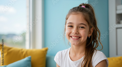 Free Download Brunette girl with ponytail and pink hair tie smiling in bright living room with turquoise walls and yellow pillows. Cheerful home portrait for Children's Day and interior design campaigns - GFXLoad