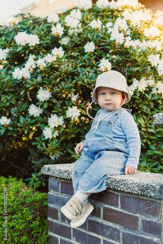 Obraz na płótnie A happy child sits on a sturdy brick wall in a blooming garden, embodying the joy of childhood. Dressed in cute overalls, the child radiates happiness and brings cheerful energy to the scene w salonie