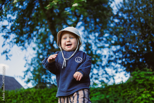 A cheerful toddler plays outside in nature, dressed comfortably, smiling brightly under the warm sun, exploring surroundings and seeking adventures filled with pure happiness and laughter