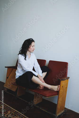 Obraz na płótnie A contemplative woman in casual attire sits on vintage seating in a minimalist space, radiating elegance. Her relaxed pose and the serene atmosphere create a captivating scene for photography w salonie