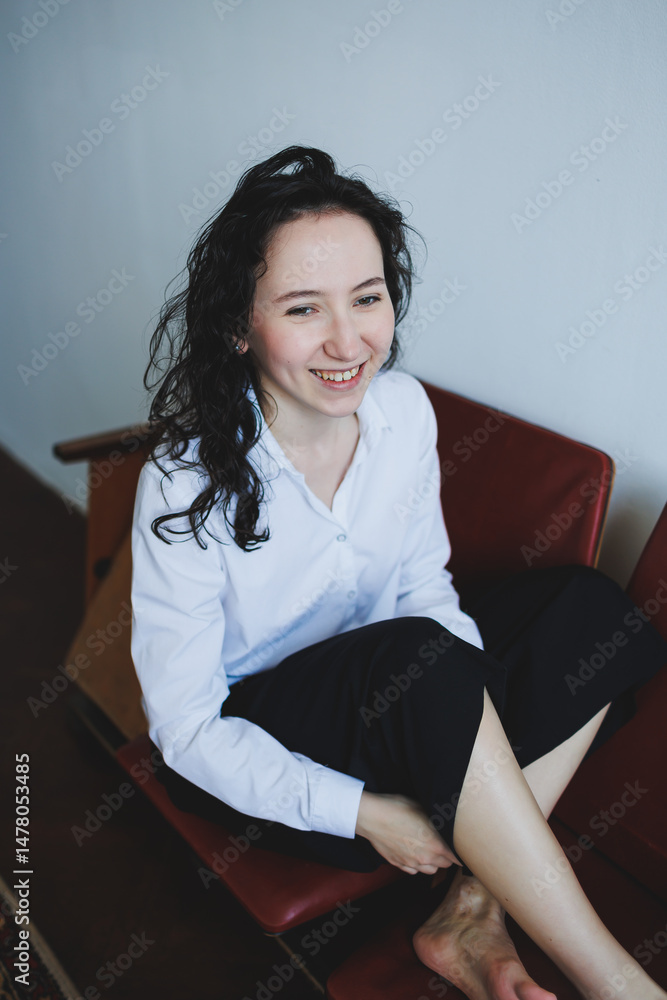 A young woman sits on a modern couch, dressed casually yet elegantly, smiling warmly in a minimalist indoor setting that reflects happiness, serenity, and a sense of peace and contentment