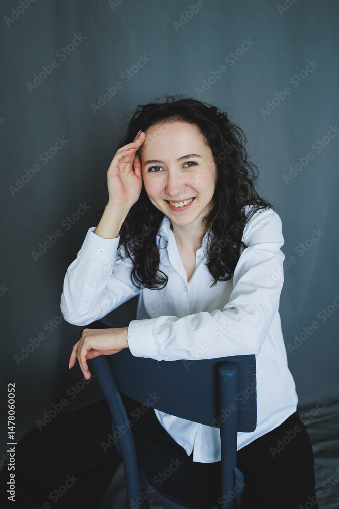 A young woman dressed in a white shirt is comfortably sitting on a chair, radiating a cheerful expression that perfectly captures a beautiful moment filled with joy, peace, and relaxation