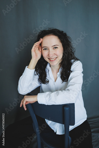 Obraz na płótnie A young woman dressed in a white shirt is comfortably sitting on a chair, radiating a cheerful expression that perfectly captures a beautiful moment filled with joy, peace, and relaxation w salonie