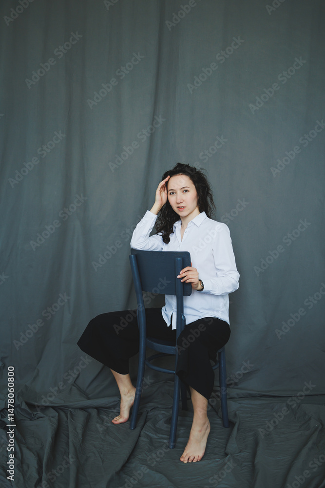 A Contemplative Portrait Featuring a Young Woman Seated Comfortably on a Chair, Set Against a Soft Gray Backdrop, Exuding a Mood of Reflection and Introspection that Engages the Viewer Interest