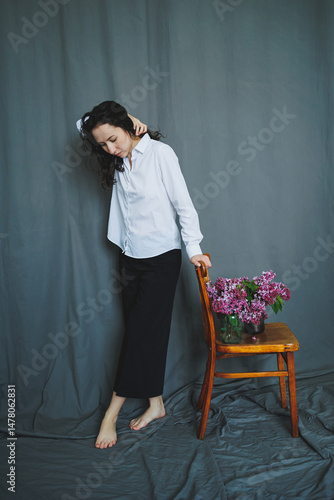 Obraz na płótnie An Elegant Portrait of a Woman Dressed in a Crisp White Shirt and Classic Black Pants, Gracefully Holding Fresh Lilacs while Seated on a Vintage Wooden Chair, Creating a Beautiful Atmosphere w salonie