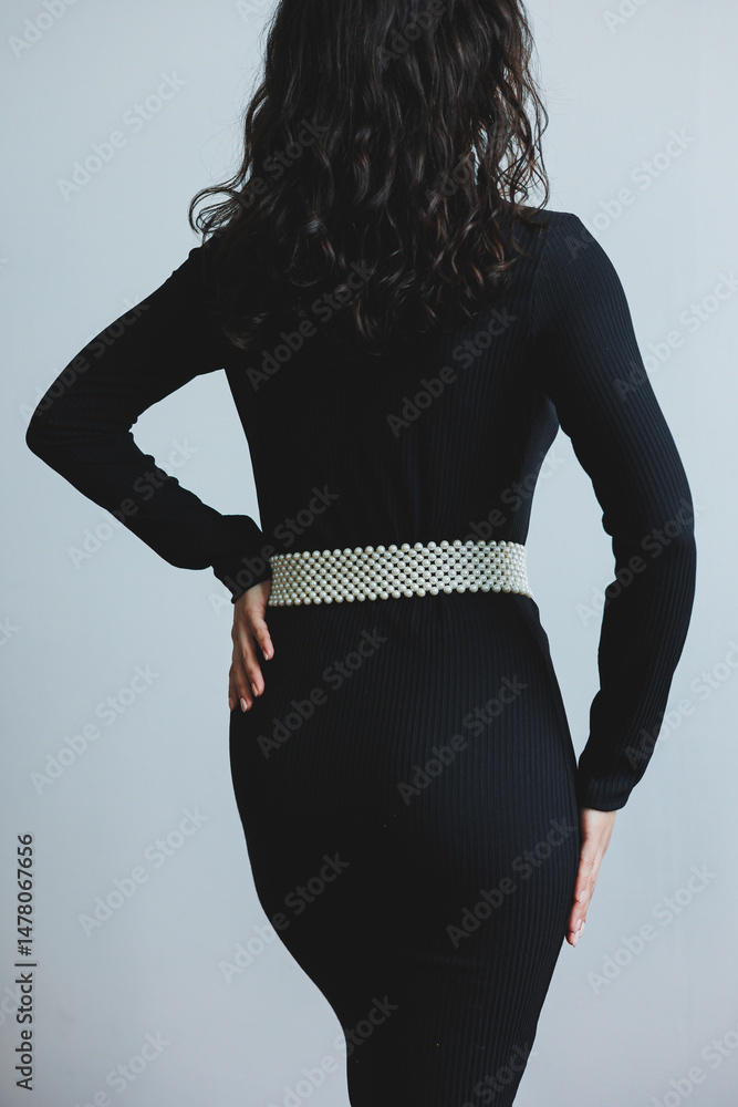 An elegant woman striking a captivating pose while wearing a sophisticated black dress that features a statement belt. She stands against a minimalist background that enhances her beauty and style