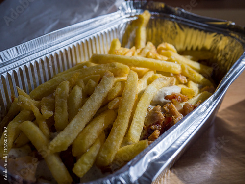 French fries piled on a kebab drizzled with sauce, all nestled in an aluminum takeaway box resting on a wooden table, creating a tempting fast food meal