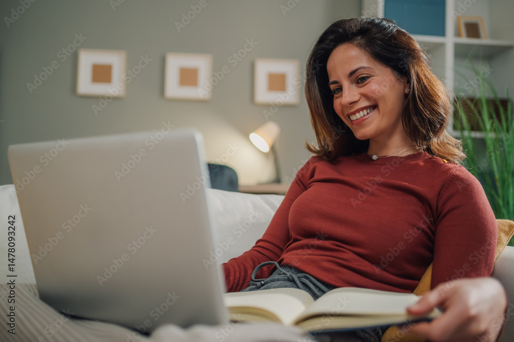 Smiling woman using laptop and reading book on sofa at home Smiling woman using laptop and reading book on sofa at home