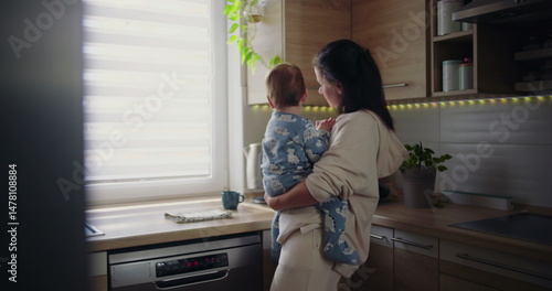 Mother holding baby in kitchen while pouring water into cup, multitasking parenting moment during casual morning routine, warm and nurturing family scene