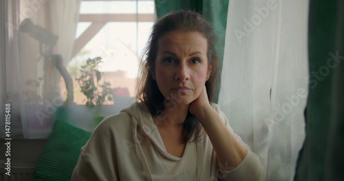Woman looking directly at the camera with a calm and composed expression, sitting near a window in a cozy home interior with plants and green curtains