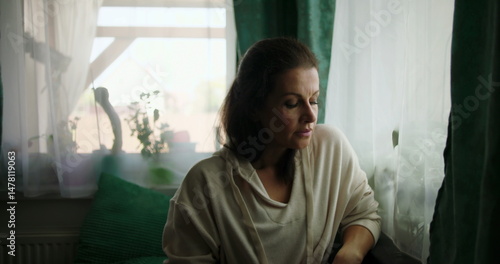 Woman sitting on a sofa, hands clasped on her lap, gazing out of a window with a thoughtful expression, surrounded by cozy home interior and soft decor