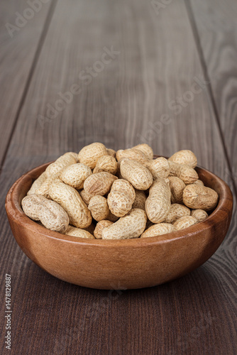 Close-up photo of the wooden bowl full of roasted unshelled peanuts on the wooden table background with copy space.