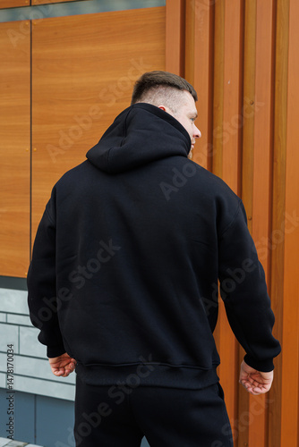 Obraz na płótnie An athletic man in a sleek black hoodie confidently shows off his fit physique in an outdoor setting with modern architecture, blending fitness and fashion into an inspiring lifestyle w salonie