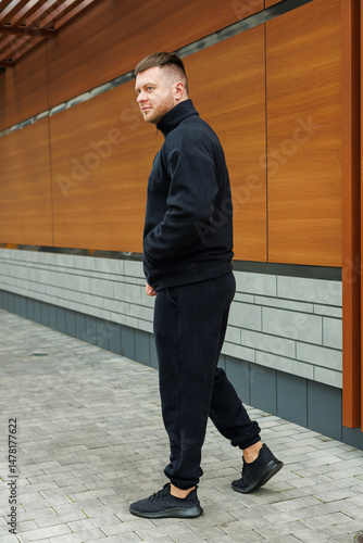 A stylish young man poses outdoors in a black tracksuit against striking modern architecture, showcasing his contemporary style and vibrant energy, embodying modern fashion and elegance