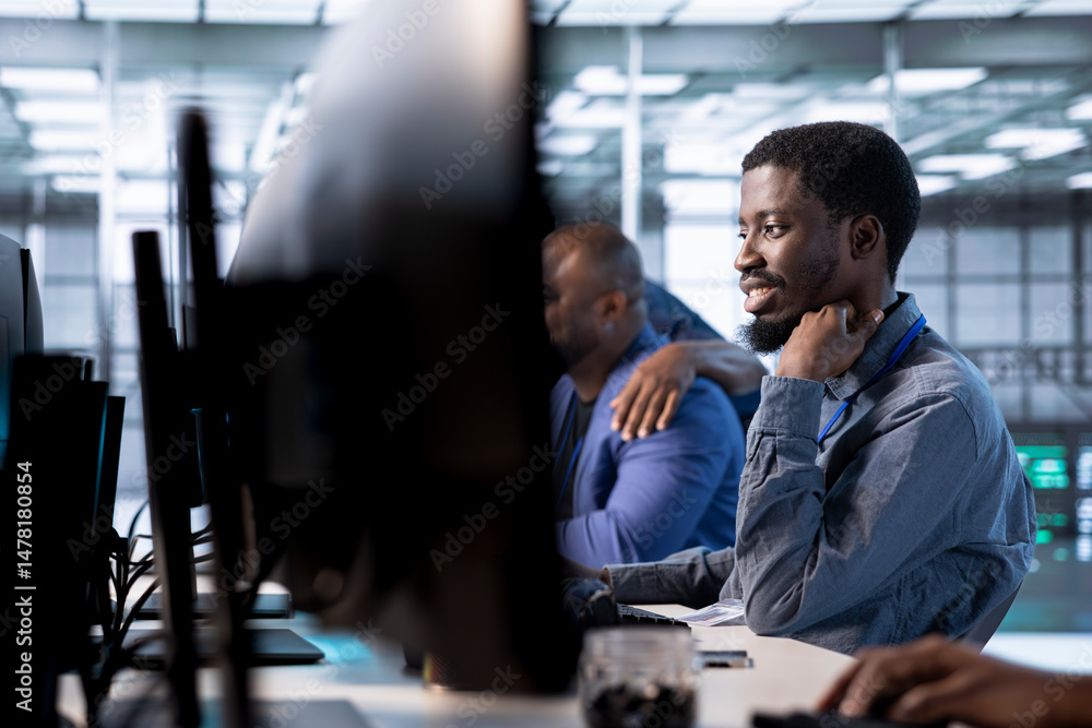 Fototapeta premium Smiling engineer in data center next to colleagues updating systems to keeps performance up and protect servers from software vulnerabilities. Coworkers looking for errors causing servers to slow down