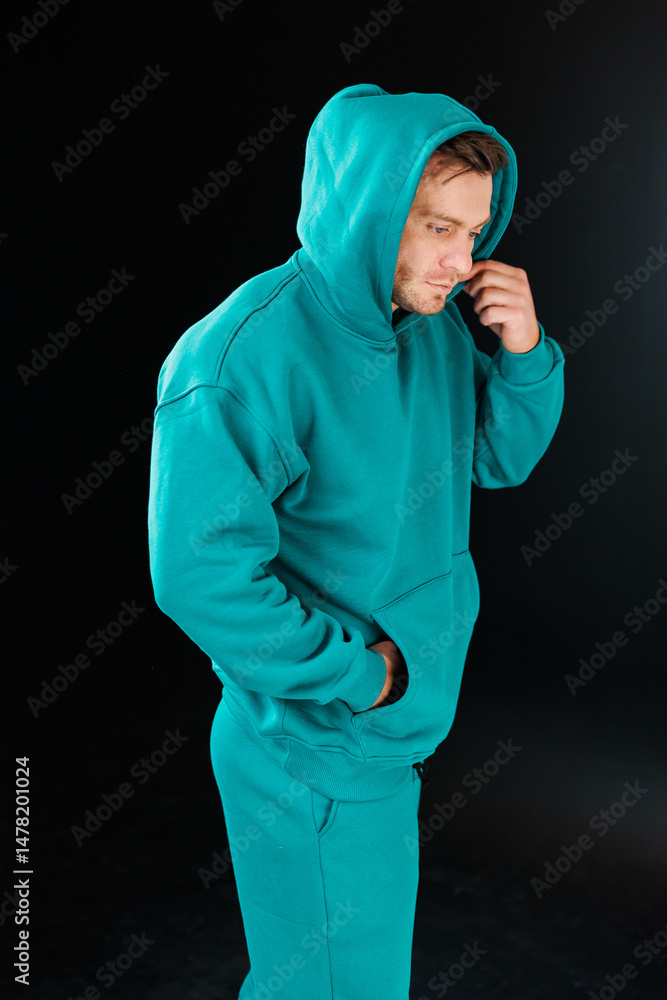 A fashionable man wearing an aqua hooded sweatshirt strikes a confident pose, exuding style as he stands against a dark, moody background that enhances the visual impact of the scene