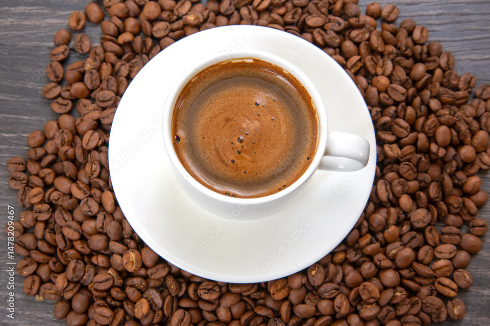 An overhead view showcasing a small white cup of espresso with a creamy foam, set on a saucer and encircled by a rustic backdrop of dark roasted coffee beans. An overhead view showcasing a small white cup of espresso with a creamy foam, set on a saucer and encircled by a rustic backdrop of dark roasted coffee beans.