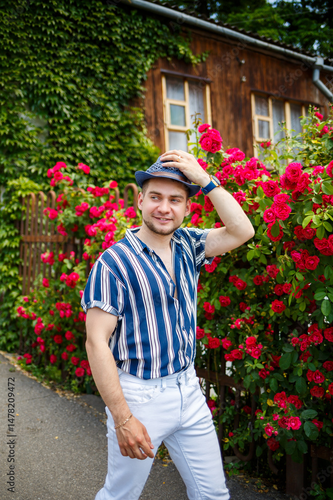 In a vibrant garden of blooming roses, a stylish young man in a blue and white striped shirt poses confidently, showcasing his cheerful and friendly personality with a bright smile