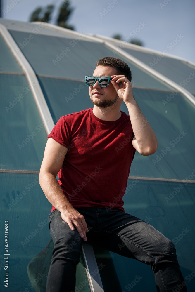 A stylish young man poses confidently in trendy sunglasses, set against modern architecture that reflects the vibrant spirit of contemporary urban lifestyle, showcasing a relaxed yet dynamic vibe