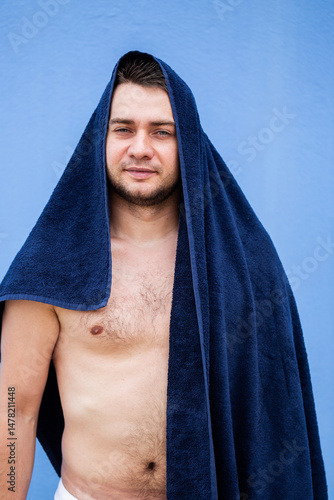 Obraz na płótnie A young man draped in a towel with a calm and serene expression, positioned against a soft and soothing blue background, embodying relaxation and confidence in his casual beachwear attire w salonie